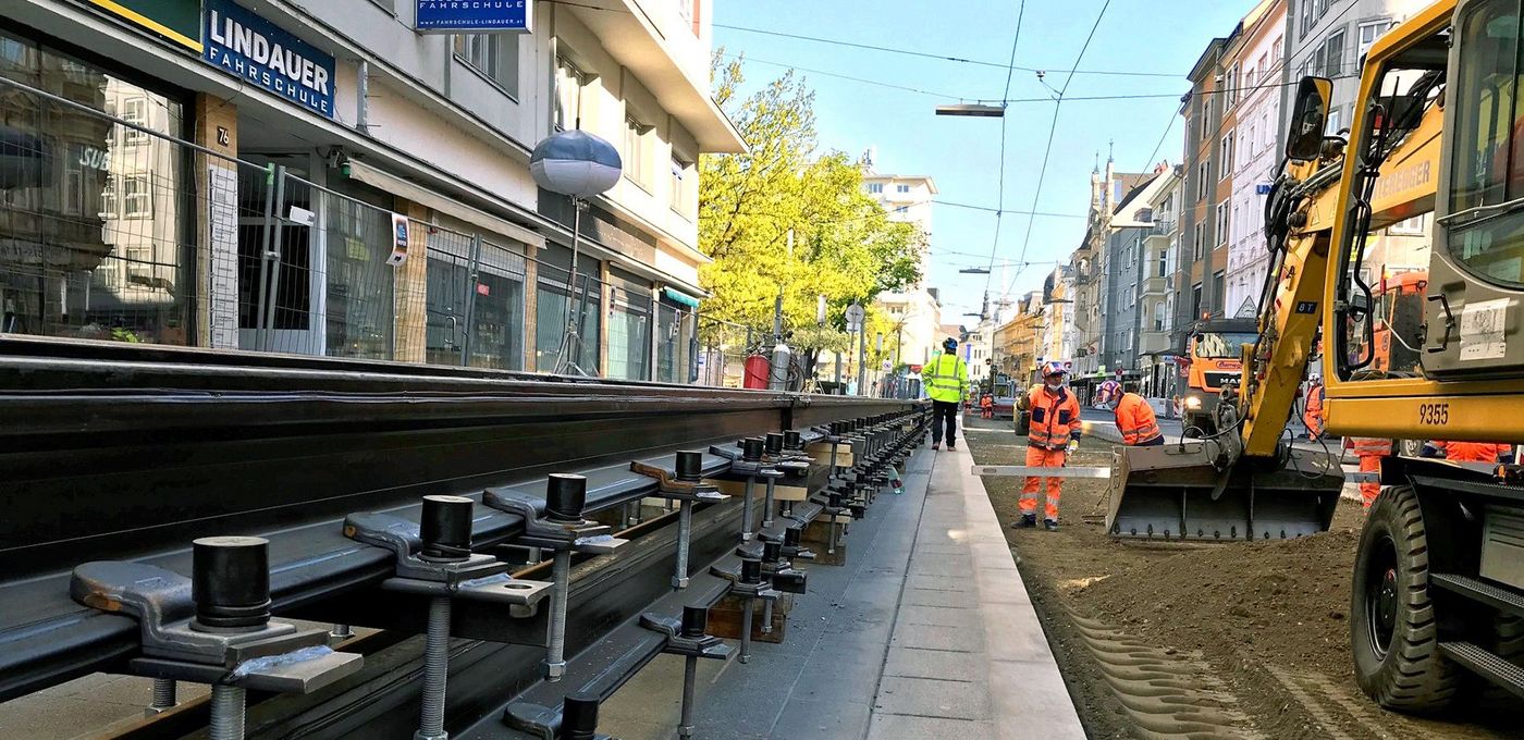 Foto: Bagger und Bauarbeiter beim Stabilisieren des Erdreichs in Fluchtperspektive einer Einkaufsstraße, links der vorbereitete neue Gleiskörper 