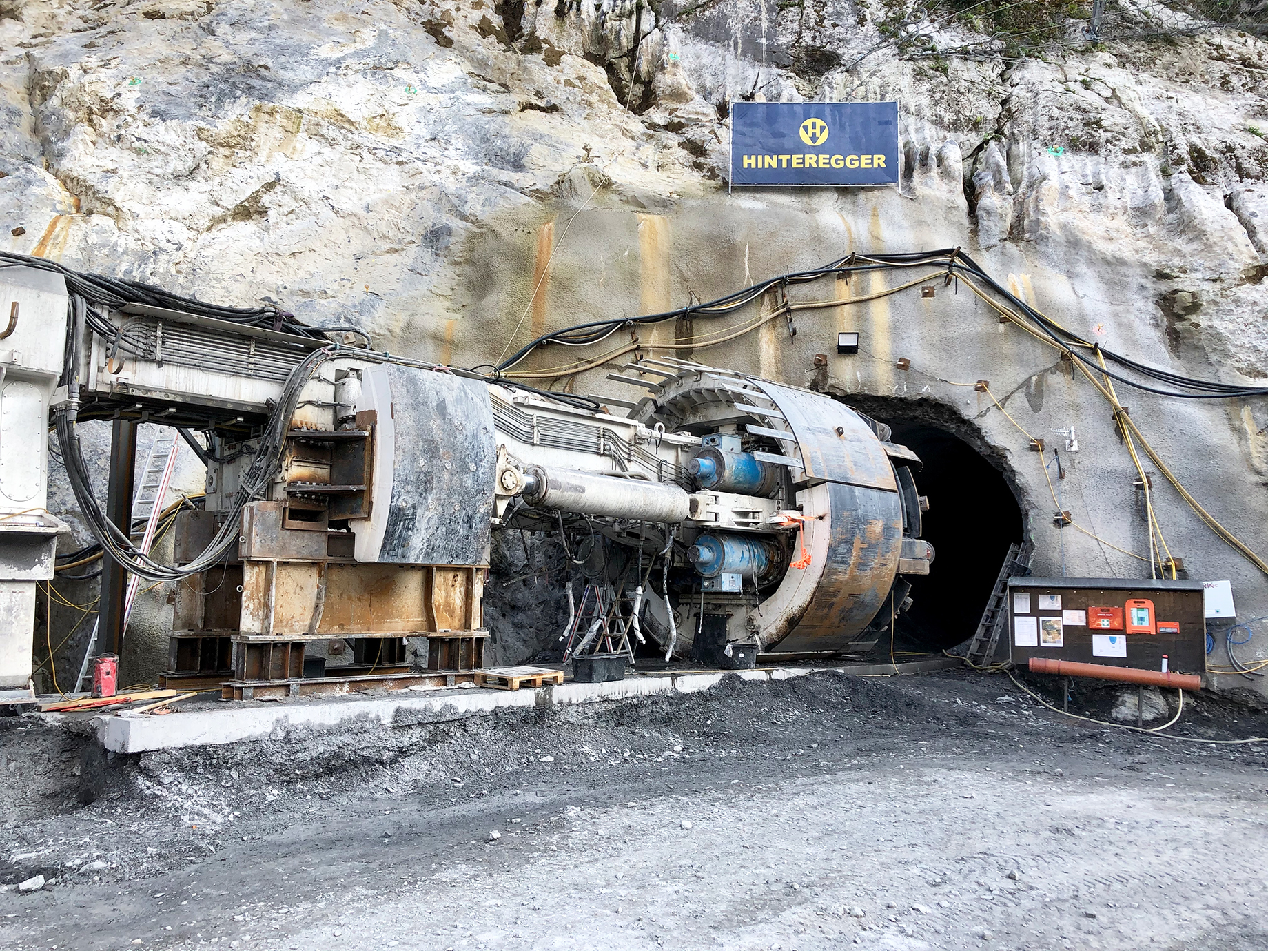 Foto: Tunnelbohrmaschine unmittelbar vor einem von Kabeln umgebenen Stolleneingang im Felsmassiv, darüber ein Hinteregger-Transparent, daneben eine Anschlagtafel mit verschiedenen Aushängen