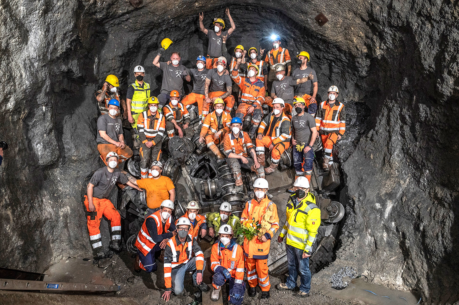 Foto: Tunnelbauteam aus rund 30 Personen mit Schutzhelmen und MNS-Masken vor und auf einer TBM inmitten von Felsen